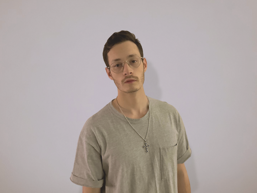 Proppa press shot in a grey shirt, thin silver chain, and glasses.